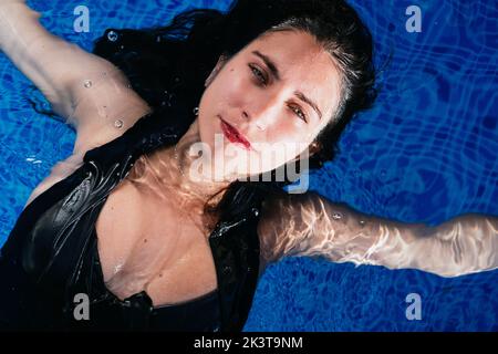 Au-dessus de la jeune femme détendue dans le bikini flottant dans l'eau de cristal de la piscine bleue tout en appréciant les vacances d'été Banque D'Images