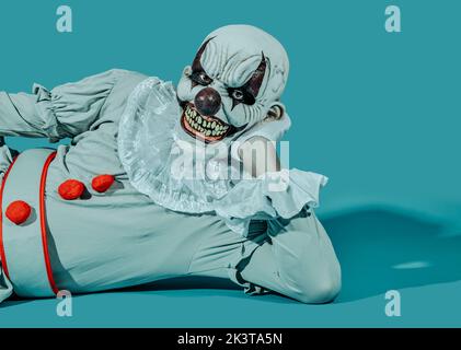 un clown mal chauve crépeux, avec un sourire menaçant, portant un costume gris avec une robe blanche, est allongé sur son côté, penché sur un bras, sur un backgro bleu Banque D'Images