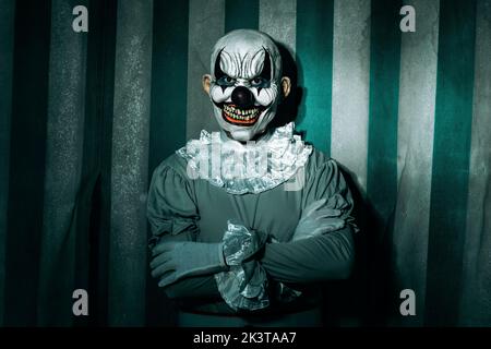 gros plan d'un clown maléfique et éhontée, debout devant une tente de cirque sale et ancienne, portant un costume gris avec une ruff blanche, et mettant en vedette à l'o Banque D'Images