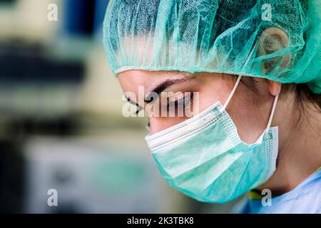 Vue de côté de récolte d'une femme médecin sérieuse et concentrée travaillant dans un masque vert clair et une coiffe chirurgicale en polyéthylène pendant l'opération à l'hôpital Banque D'Images