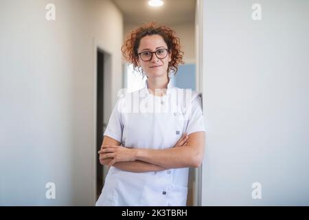 Jeune femme thérapeute positive en uniforme blanc et lunettes souriant et regardant la caméra tout en se tenant dans le couloir de la clinique moderne Banque D'Images