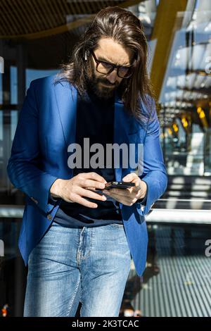 Homme d'affaires moderne aux cheveux longs habillé dans une veste élégante et jeans naviguant sur Internet sur un téléphone portable tout en étant debout avec une valise à côté du mur en verre i Banque D'Images