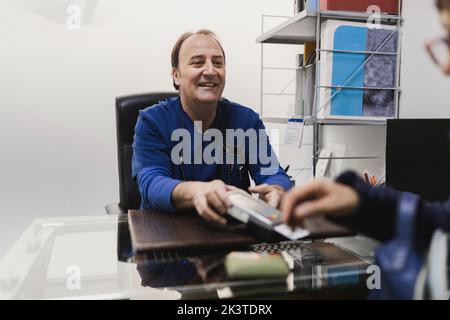 Positif adulte homme médecin en uniforme donnant le terminal de paiement avec carte en plastique au client de récolte à la table en vétérinaire Banque D'Images