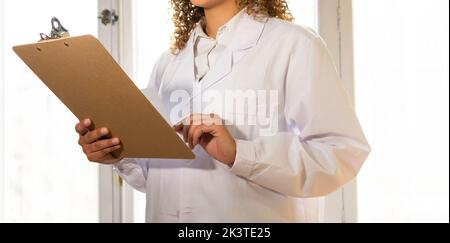 Anonyme ethnique féminine medic en uniforme regardant le presse-papiers tout en travaillant contre la fenêtre dans la clinique Banque D'Images