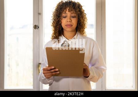 Femme ethnique attentive medic en uniforme regardant la planchette à pince tout en travaillant contre la fenêtre dans la clinique Banque D'Images