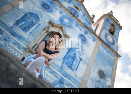 Faible angle de jeune contenu touristique femelle reposant sur les escaliers contre l'extérieur de l'ancienne église avec la murale tout en regardant loin dans Agueda Portugal Banque D'Images