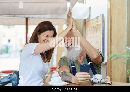 Bonne femme et petit-père tatoué barbu avec téléphone portable donnant cinq hauts en riant à table avec café dans le café Banque D'Images