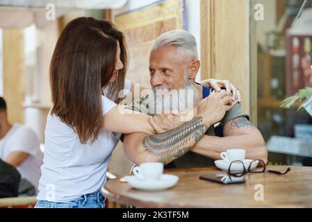 Femme souriante adulte qui se fait embrasser et poser avec un grand-père chic et hippster sur un téléphone portable tout en regardant les uns les autres à une table de café Banque D'Images