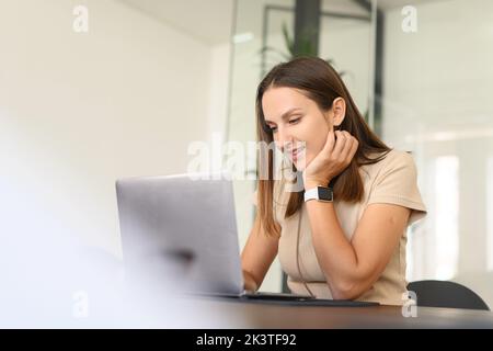 Portrait d'une jeune femme caucasienne assise au bureau avec un ordinateur portable, en tapant un e-mail, en envoyant un message à un ami, en discutant, datant en ligne avec un sourire agréable, concept de relations à distance Banque D'Images