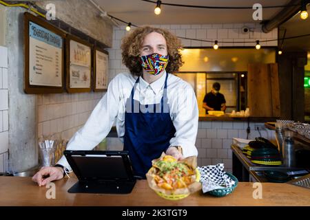 Jeune homme positif serveur dans le masque facial et le tablier debout au comptoir de la cafétéria et regardant la caméra Banque D'Images