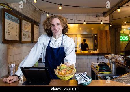 Positif jeune homme serveur heureux dans un tablier debout au comptoir de la cafétéria et regardant la caméra Banque D'Images