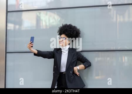 Joyeuse architecte afro-américain dans une tenue élégante debout près d'un mur de verre et prenant autoportrait sur smartphone en ville Banque D'Images