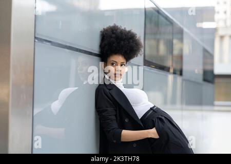 Vue latérale de l'architecte noir féminin en costume tendance et avec la coiffure afro penchée sur le mur de verre du bâtiment tout en se tenant avec les mains dans les poches et Banque D'Images