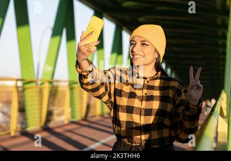 Jeune femme jeune femme ethnique confiante avec de longs cheveux foncés dans une tenue élégante et un chapeau souriant et montrant deux doigts de geste tout en prenant le selfie Banque D'Images