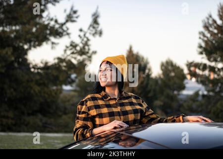 Jeune touriste ethnique auto-assurée avec de longs cheveux foncés dans une chemise et un chapeau debout près d'une voiture garée sur une pelouse herbacée dans la campagne et regardant loin Banque D'Images