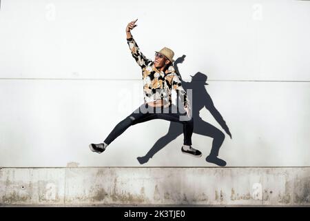 Sortie d'un homme afro-américain dans une tenue tendance souriant et prenant le selfie tout en sautant près du mur grundy sur la rue de la ville Banque D'Images
