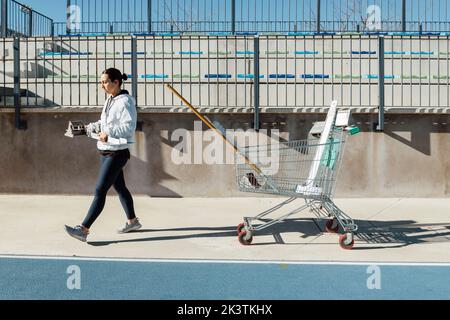 Vue latérale d'une jeune athlète féminine transportant des blocs de départ tout en marchant près d'un chariot avec de l'équipement avant l'entraînement sur piste et terrain le jour ensoleillé sur le stade Banque D'Images