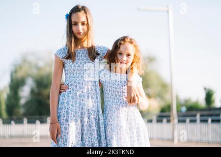 Deux jolies filles dans des robes à motifs similaires s'embrassant et regardant l'appareil photo tout en se tenant sur un arrière-plan flou sur la rue suburbaine sur su Banque D'Images