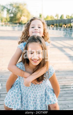 Jolie fille en robe à motifs souriant et portant une jeune sœur dans le dos tout en se tenant sur un fond flou de la rue de banlieue par beau temps Banque D'Images