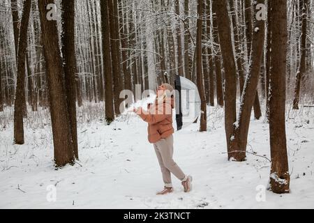 Une jeune femme se tient dans une forêt de pins avec un parapluie transparent, en collant sa langue pour attraper des flocons de neige. Banque D'Images