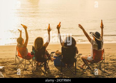 Vue arrière d'un groupe de jeunes amis gaies assis sur des chaises et des bouteilles de bière tout en se rassemblant sur une plage de sable en soirée d'été Banque D'Images