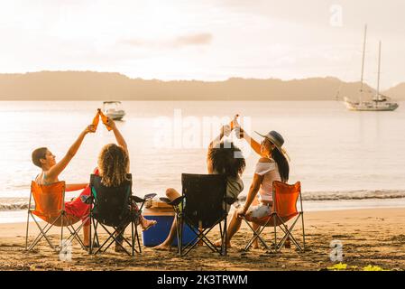 Vue arrière d'un groupe de jeunes amis gaies assis sur des chaises et des bouteilles de bière tout en se rassemblant sur une plage de sable en soirée d'été Banque D'Images