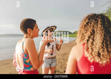 Groupe de jeunes gens de l'ethnie gaie bavardant amicaux et riant tout en mangeant de la pizza et de boire de la bière ensemble sur le sable de mer au Costa Rica Banque D'Images