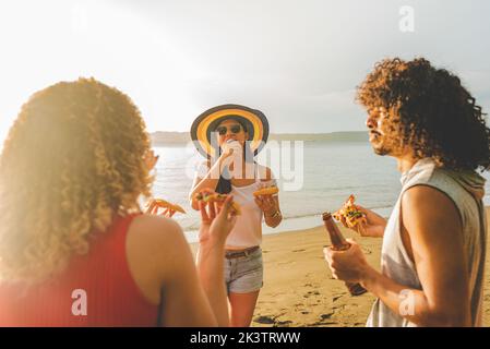 Groupe de jeunes gens de l'ethnie gaie bavardant amicaux et riant tout en mangeant de la pizza et de boire de la bière ensemble sur le sable de mer au Costa Rica Banque D'Images