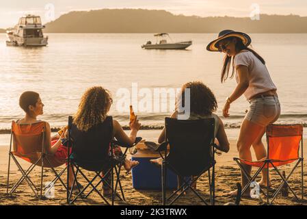 Vue arrière d'un groupe de jeunes amis gaies assis sur des chaises et des bouteilles de bière tout en se rassemblant sur une plage de sable en soirée d'été Banque D'Images