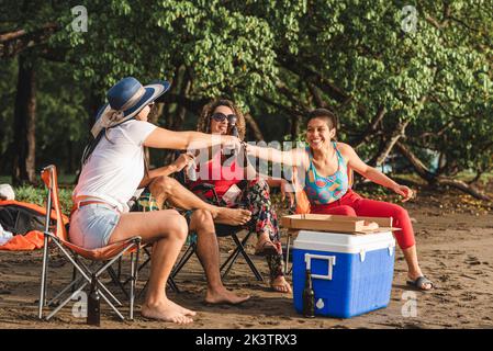 Groupe de jeunes amis gais assis sur des chaises et des bouteilles de bière tout en se rassemblant sur la plage de sable en soirée d'été au Costa Rica Banque D'Images