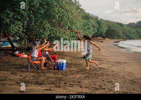 Jeune homme ethnique transportant une boîte à pizza et courant vers un groupe d'amis heureux en profitant d'un pique-nique d'été sur la plage de sable au Costa Rica Banque D'Images