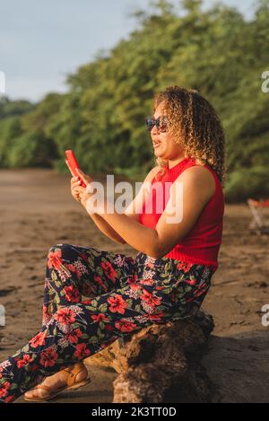 Vue latérale une femme ethnique aux cheveux bouclés dans une tenue d'été colorée et lumineuse et des lunettes de soleil assis sur une plage de sable, parcourant un téléphone portable en été Banque D'Images