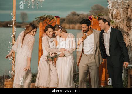 Jeunes souriants dans des costumes embrassant près de belles dames joyeuses dans des robes avec bouquet de fleurs près de décorations par temps nuageux Banque D'Images