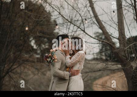 Vue latérale d'un jeune garçon en costume embrassant et embrassant avec une dame en robe de mariage avec bouquet de fleurs à la main près des arbres sur fond flou Banque D'Images