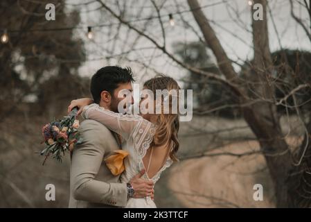 Vue latérale d'un jeune garçon en costume avec dame en robe de mariage avec bouquet de fleurs à la main près des arbres sur fond flou Banque D'Images