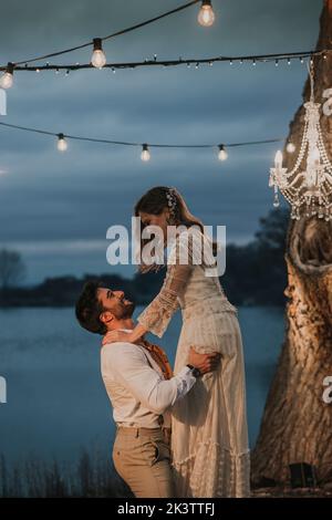Jeune homme soulevant une mariée gaie sous un arbre et des lustres près du lac la nuit Banque D'Images