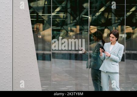 Femme entrepreneur élégante dans des vêtements élégants et décontractés, debout près d'un bâtiment en verre en ville, tout en utilisant des écouteurs et en naviguant sur un smartphone Banque D'Images
