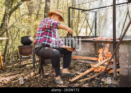 Vue latérale d'un homme argentin versant de l'eau chaude de la bouilloire dans la gourde de calabash contre le rack au-dessus du feu brûlant dans la campagne Banque D'Images