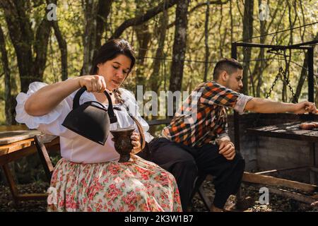 Une femme Argentine verse de l'eau chaude de la bouilloire dans une gourde de calabash contre un fils de sexe masculin qui fait griller de la viande au-dessus du feu dans le camping Banque D'Images