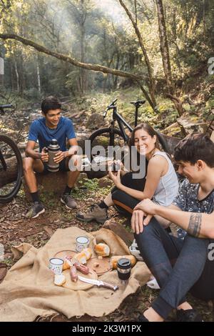 Divers cyclistes assis sur le sol près de la tente et ayant pique-nique tout en randonnée ensemble dans la forêt Banque D'Images