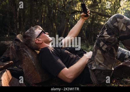 Vue latérale d'un touriste de sexe masculin d'âge moyen en lunettes de soleil prenant un autoportrait sur un téléphone portable tout en se penchant sur la pierre dans le camping Banque D'Images