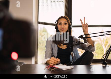 Hôte radio femelle dans un casque assis à la table avec microphone regardant la caméra faisant le signe V pendant l'enregistrement de la vidéo Banque D'Images