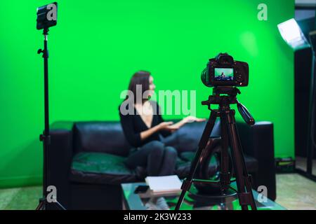 Femme blogueuse souriante assise sur un canapé en cuir et pointant vers l'extérieur tout en enregistrant la vidéo sur fond de chrominkey Banque D'Images