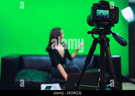 Femme blogueuse souriante assise sur un canapé en cuir et pointant vers l'extérieur tout en enregistrant la vidéo sur fond de chrominkey Banque D'Images