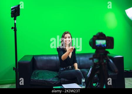 Femme blogueuse souriante assise sur un canapé en cuir et pointant vers l'appareil photo sur un trépied tout en enregistrant la vidéo sur fond chromakey Banque D'Images