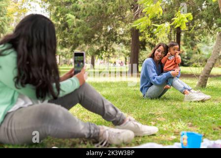 Jeune femme qui prend des photos sur le téléphone portable d'une maman gaie qui élève son bébé tout en étant assise sur la pelouse dans le parc Banque D'Images