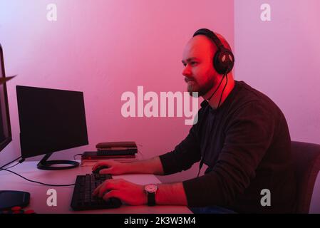 Un homme à barbe chauve portant un casque est assis et travaille à l'ordinateur. Prise de vue en lumière rouge. Banque D'Images