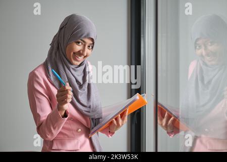 Portrait d'une femme d'affaires souriante portant le hijab au bureau près d'une fenêtre. Banque D'Images