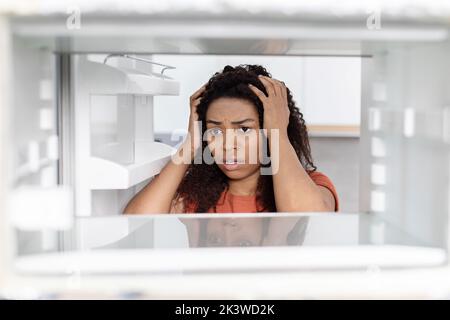 Triste choquée femme noire millénaire tient la tête, regardant dans le réfrigérateur vide, souffrant de la famine Banque D'Images