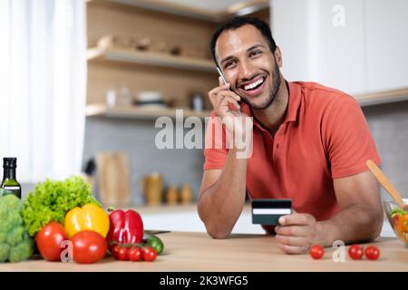 Un homme noir millénaire souriant à table avec des légumes biologiques utilise une carte de crédit et les appels par téléphone Banque D'Images